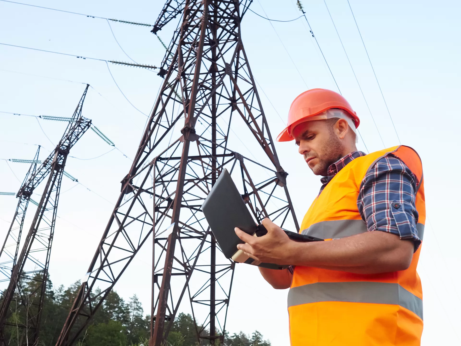 Worker in orange vest using laptop near power lines, showcasing PMC Wireless technology solutions for utilities.