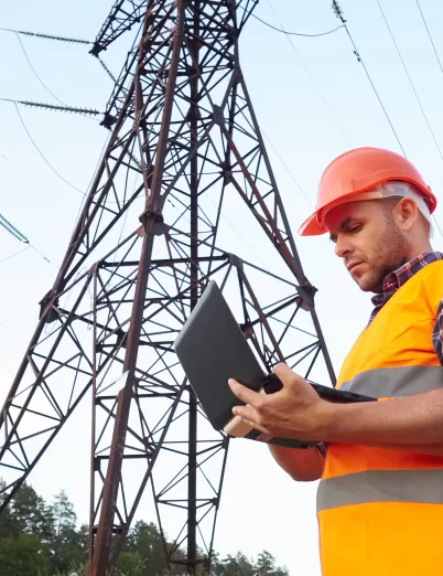 Worker in orange vest using laptop near power lines, showcasing PMC Wireless technology solutions for utilities.