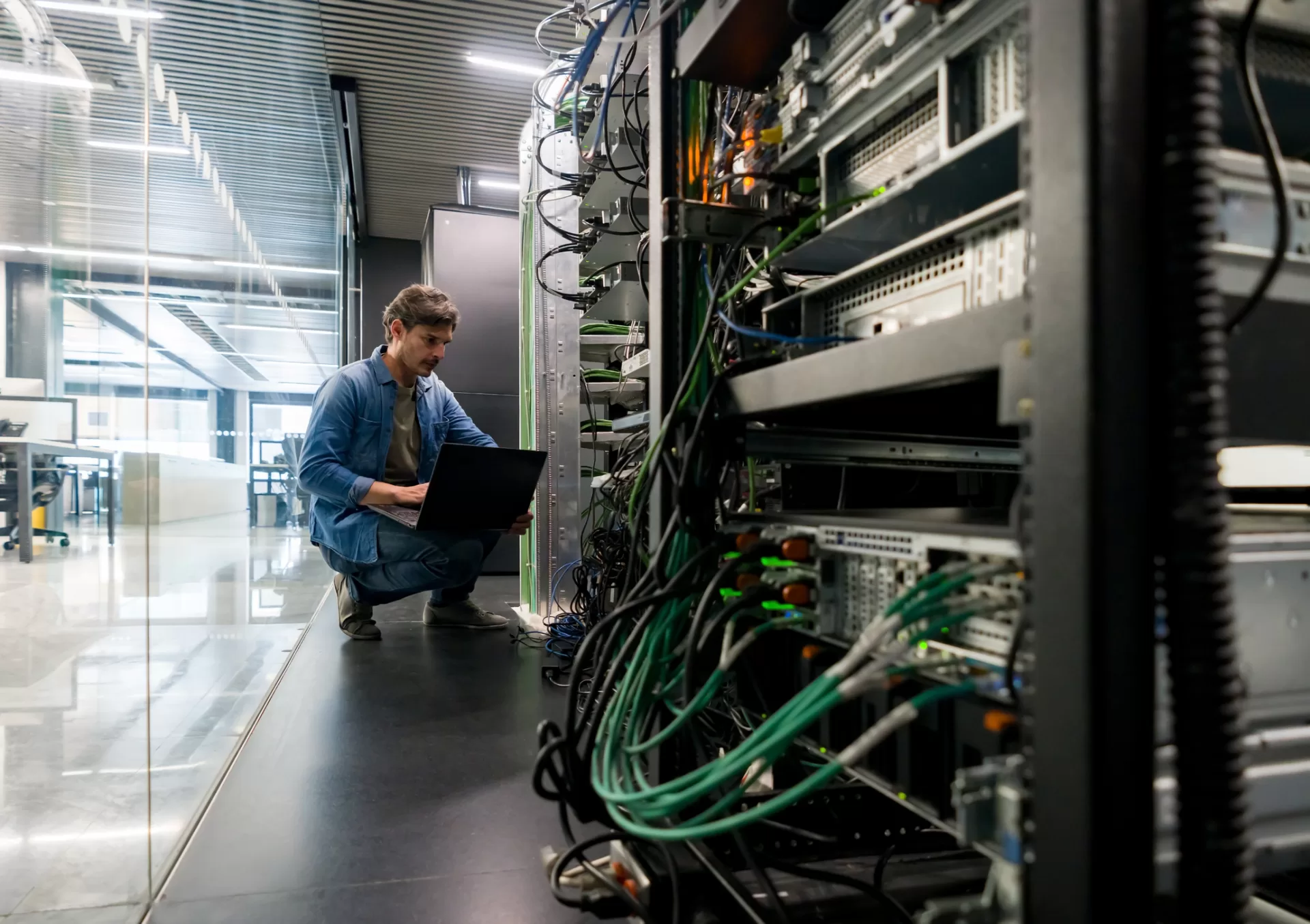 Man configuring network settings on a laptop beside a server rack in a modern PMC Wireless office.