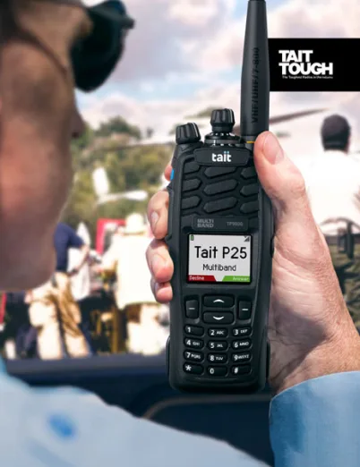Person holding Tait TP-9900 multi-band portable radio displaying "Tait P25" on screen, showcasing its rugged design and functionality in a public safety context.